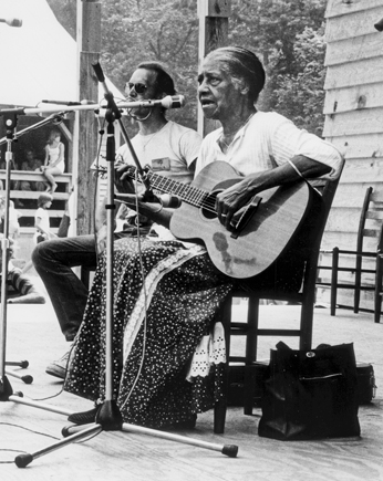 Elizabeth Cotten sings into a microphone. She is wearing a long skirt and white shirt. She is strumming a guitar. Her hair is pulled back to the nape of her neck. She is older.